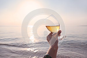 Pov photo of woman hand holding glass of white wine over sea sunset on the beach