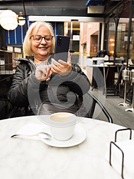 pov senior woman laughing sending selfie on cell phone with coffees already finished