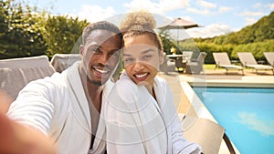 POV Shot Of Couple In Robes Outdoors By Pool On Spa Day Posing For Selfie On Mobile Phone