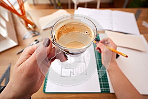 POV Shot Of Female Architect Holding Cup Of Coffee Working In Office On Plans For New Building