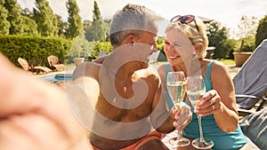 POV Shot Of Senior Couple On Holiday Posing For Selfie Drinking Champagne Relaxing By Swimming Pool 