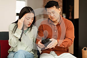 Povertry and debt concept. Unhappy asian spouses having financial problems, looking at empty wallet, sitting on couch