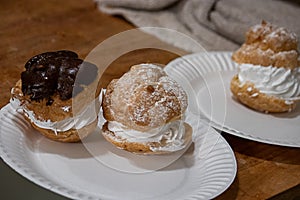 Powdered sugar and shiny chocolate glased cream puffs with a swirl of white whipped cream on a paper plate