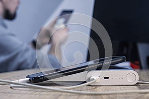 Power bank with a phone and a cable on the background of a man sitting at the monitor. A portable charger charges a smartphone on