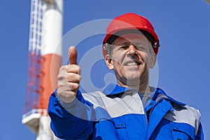 Power Plant Worker Giving Thumbs Up Against Industrial Chimney and Blue Sky Background