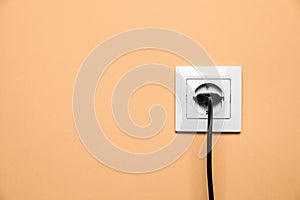 Power socket with inserted plug on pale orange wall, space for text. Electrical supply