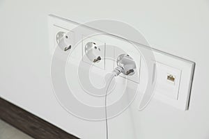 Power sockets with inserted plug on white wall indoors, closeup. Electrical supply