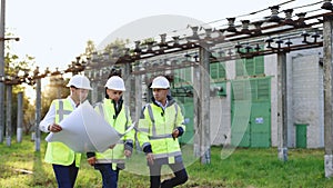 Power specialists are planning a new project outdoors. Three engineers walk near power lines in the high voltage power