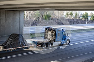 Powerful big rig blue semi truck tractor transporting covered cargo on step down semi trailer running on the wide highway road