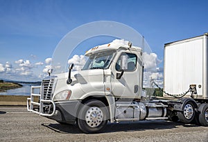 Big rig white day cab semi truck with grille guard transporting cargo in dry van semi trailer running on the road at sunny day