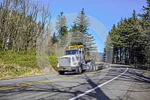 Powerful big rig white semi truck with oversize load sign on the roof transporting empty step down semi trailer for carry heavy-