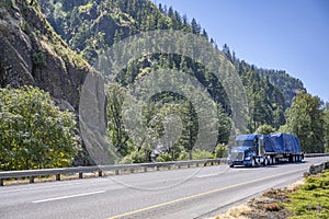 Powerful blue big rig semi truck transporting heavy duty cargo covered with tarp on flat bed semi trailer driving on the highway