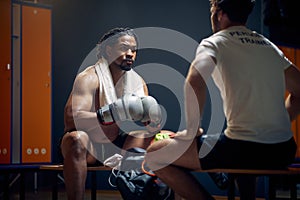 Powerful boxer in sweats resting on bench after workout, talking with his coach, getting consultation