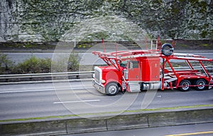Powerful classic big rig red car hauler semi truck running with empty semi trailer on the divided road with ivy covered wall