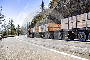 Powerful classic big rig semi truck tractor transporting fastened lumber wood on flat bed semi trailers running on the mountain