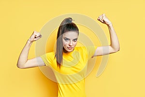 Powerful confident young woman showing arms muscles on yellow background