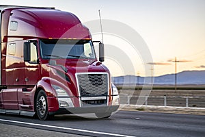 Powerful dark red bonnet big rig semi truck transporting cargo in dry van semi trailer driving on the one way highway road at