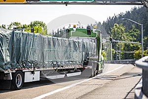 Powerful green big rig semi truck with covered cargo on step down semi trailer running on the city street with traffic light on