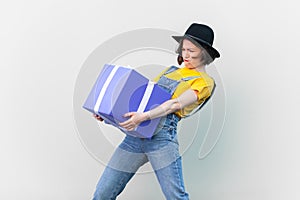 Powerful hipster woman in blue denim overalls, holding very heavy present box, making effort.