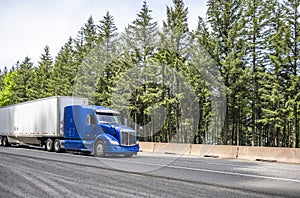 Powerful industrial long haul blue big rig semi truck transporting goods in dry van semi trailer running on the wide highway road