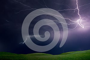 Powerful lightnings in dark stormy sky above green grass hills