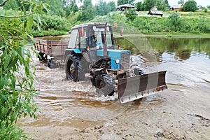 A powerful, passable, blue tractor with a trailer moves along the river. Crossing the river. Summer. The photograph is horizontal