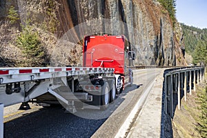 Powerful red big rig classic semi truck tractor transporting empty flat bed semi trailer running on the narrow mountain road with