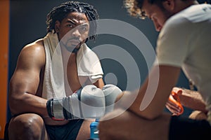 Powerful strong young boxing player with his coach, getting prepared for workout, sitting on bench face to face