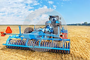 A powerful universal blue harrow on an agricultural tractor against the background of a harvested yellow wheat field