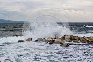 Powerful wave in sea crashing against rock sending up sprays of white foam in sky with cirrus clouds seagull above horizon line