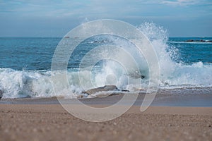 Powerful wave splashing against rocks on beach, throwing spray into the air.