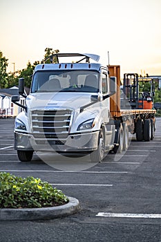 Powerful white day cab big rig semi truck with roof spoiler and flat bed semi trailer with fork lift on the back standing on the