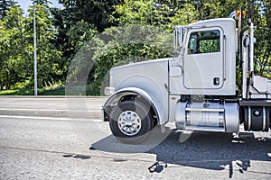 Powerful white day cab big rig semi truck tractor driving on the sunny summer highway road with green forest on the background