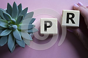 PR - Public Relations symbol. Concept word PR on wooden cubes. Businessman hand. Beautiful pink background with succulent plant.