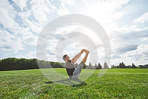 Practicing yoga. Sporty young man stretching outdoors on a green lawn in open field on a sunny morning. Yoga meditation.