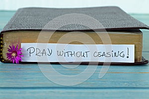 Pray without ceasing, a handwritten text on a note with a purple flower in front of a closed Holy Bible Book on wood