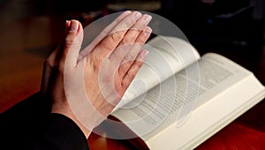 Pray to God. Female hand in prayer over a Holy Bible, wooden table, close up view