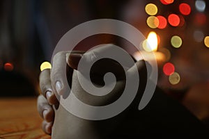 Prayer hands of a young girl on background of candle light and colorful Christmas bokeh lights. Person praying alone in silent.