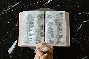 Praying hands folded in prayer to God Jesus Christ with open Holy Bible Book on dark granite background