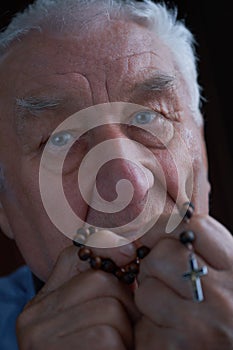 Praying old man holding a rosary beads in his hands next to his mouth.