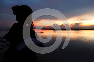 Praying silhouette of a woman on the beach against colorful morning light over sea horizon at sunrise.