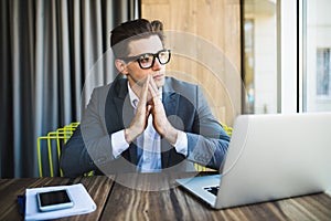 Praying for success. Front view of thoughtful young man holding hands on chin and looking at the laptop while sitting at his worki