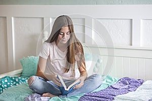 Pre-adolescent teen girl reading a book lying in bed at home. Candid indoor photo with Focus on the foreground and copy space