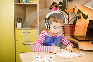 A pre-school girl with headphones is engaged at home at the table studying an educational program, writing in a notebook. Distance