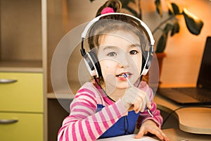 A pre-school girl with headphones is engaged at home at the table studying an educational program, writing in a notebook. Distance