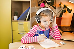 A pre-school girl with headphones is engaged at home at the table studying an educational program, writing in a notebook. Distance