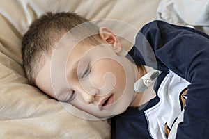 Pre-school sick boy in pyjama lying in bed with a digital thermometer