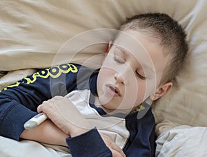 Pre-school sick boy in pyjama lying in bed with a digital thermometer in hand