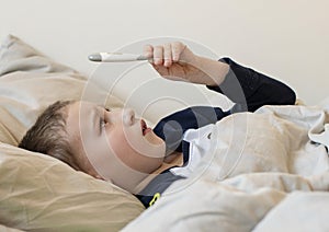 Pre-school sick boy in pyjama lying in bed and looking at digital thermometer