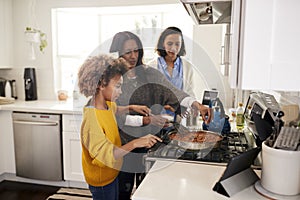 Pre-teen African American  girl standing at the hob in the kitchen preparing food with her grandmother and mother, selective focus